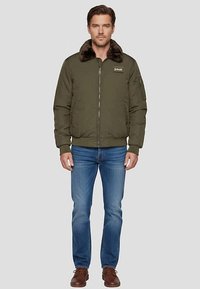 Olijfgroene bomberjack met bontkraag, ritssluiting, zijzakken, geribbelde manchetten en tailleband; gedragen met een blauwe spijkerbroek en bruine schoenen.