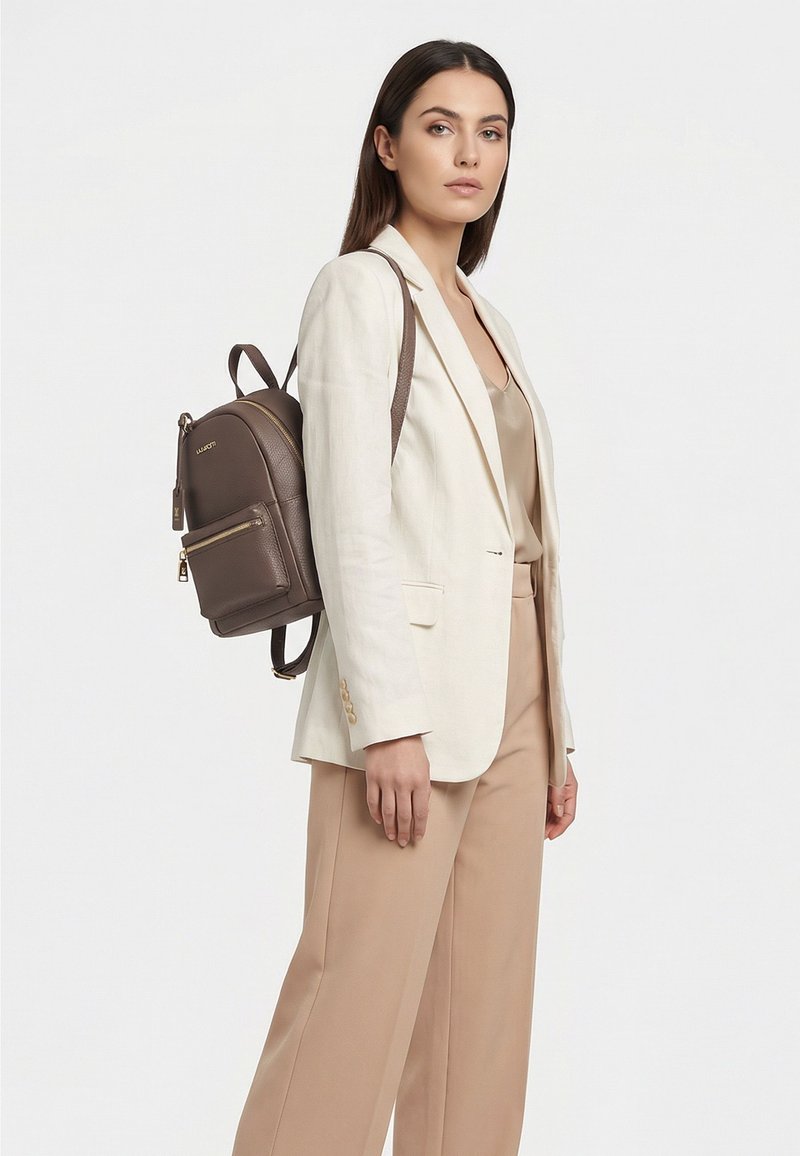Donna con pantaloni beige e blazer crema, che porta uno zaino in pelle marrone su una spalla contro uno sfondo semplice.