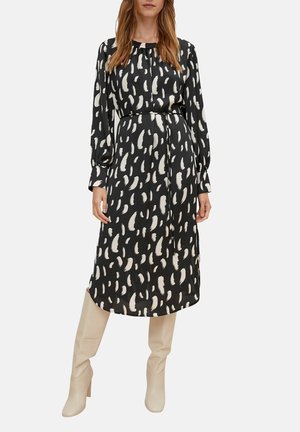 Robe noire avec un motif de coups de pinceau blancs, manches longues, taille cintrée, longueur genou, associée à des bottes beiges jusqu'aux genoux.