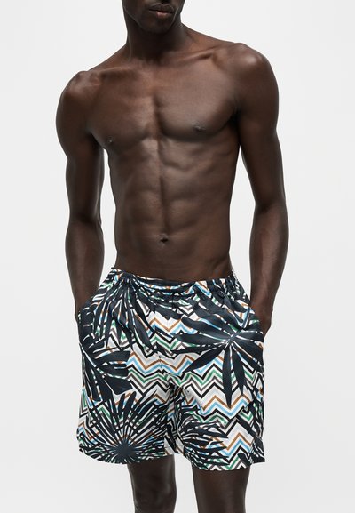 Homme torse nu à la peau foncée portant un short de bain coloré avec des motifs de feuilles de palmier et de zigzag, mains dans les poches, fond neutre.