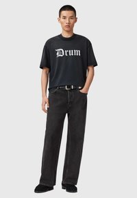 Czarna grafika na koszulce z napisem "Drum", w połączeniu z luźnymi czarnymi dżinsami i czarnym paskiem. Model stoi z jedną ręką w kieszeni.