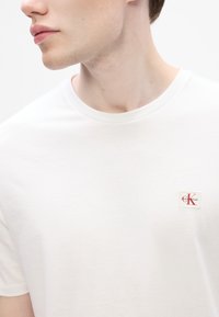 T-shirt branca de algodão com decote redondo, apresentando um pequeno patch do logótipo vermelho e branco da Calvin Klein no peito. Textura suave.