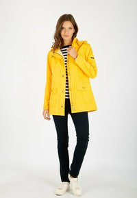 Veste imperméable jaune avec fermeture éclair et boutons-pression, deux poches avant et capuche ajustable. Portée par-dessus un haut rayé et un pantalon noir.