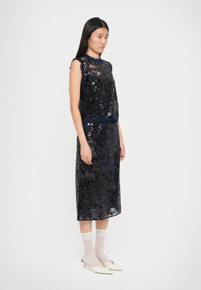 Abito senza maniche di colore navy con design floreale in paillettes, sovrapposizione trasparente e vita aderente. Abbinato a calze bianche e scarpe a punta chiare.