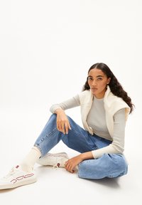 Eine Frau mit langen, lockigen Haaren sitzt im Schneidersitz und trägt einen cremfarbenen Pullover, blaue Jeans, weiße Sneakers und eine hellfarbene Weste, die ihr über die Schultern hängt.