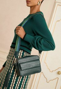 Borsa a tracolla in pelle verde intrecciata con una tracolla multicolore a righe, dotata di un pon-pon e di elementi di design eleganti.