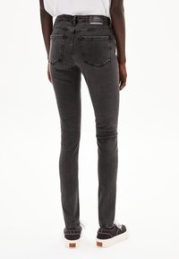 Schmal geschnittene schwarze Jeans aus einem weichen Denim-Material mit Gesäßtaschen und einem hohen Bund. Getragen mit schwarzen Schuhen mit weißen Sohlen.