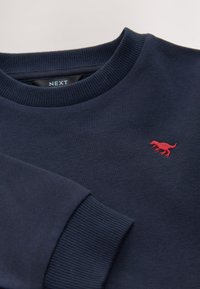 Granatowa bluza wykonana z bawełnianej mieszanki tkanin. Posiada prążkowany kołnierz i mankiety oraz małe czerwone logo dinozaura na piersi.