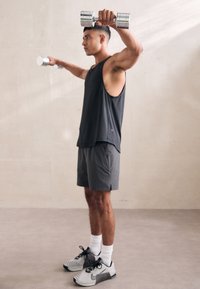 Egy férfi ujjatlan fekete trikóban és szürke rövidnadrágban ezüst súlyzókat emel. Szürke és fekete sportcipőt visel fehér zoknival.