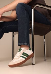 Vit sneaker med gröna ränder och detaljer, ljusgummi sula, texturerad mockatå, vilande på ett ben i mörka jeans, mot en neutral bakgrund.