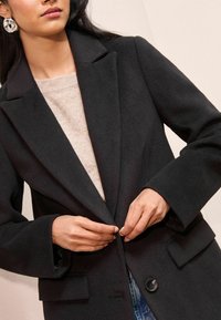Schwarzer Wollmischmantel mit eingekerbtem Kragen, frontalen Taschen und einer Knopfverschluss, kombiniert mit einem hellbeigen Pullover.
