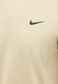 Μπεζ ύφασμα με ένα μικρό μαύρο λογότυπο Nike Swoosh κεντημένο στην πάνω περιοχή του στήθους ενός ενδύματος.
