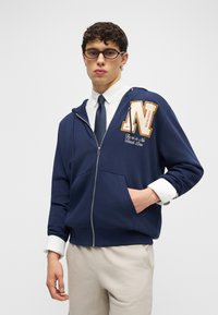 Jaunas vyras, dėvintis tamsiai mėlyną užtraukiamą džemperį su dideliu „N“ ir tekstu, baltą marškinius, tamsią kaklaraištį, šviesias kelnes ir akinius, stovintis su viena ranka kišenėje.