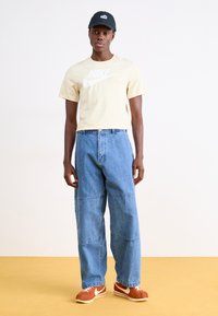 Camiseta amarilla de Nike con logo blanco, vaqueros de pierna ancha azules y zapatillas marrones de Nike. Gorra negra. Diseño simple, estilo casual.