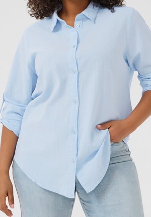 Kvinde iført lyseblå skjorte med knapper og op rullede ærmer og falmede blå jeans, med den ene hånd i lommen og krøllet hår synligt.