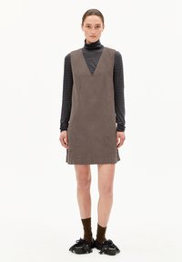 Braunes Cordkleid mit V-Ausschnitt, das eine gerade Silhouette und seitliche Nahtdetails bietet. Über einem schwarz-grau gestreiften Rollkragenpullover getragen.