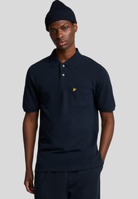 Marineblå bomulds polo-shirt med ribbet krave, korte ærmer og en påsyet lomme med et gult logo. Glat tekstur og klassisk pasform.