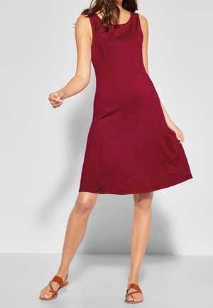 Ärmelloses burgunderrotes knielanges Kleid, getragen mit braunen flachen Sandalen, in einer lässigen stehenden Pose vor einfachem Hintergrund präsentiert.