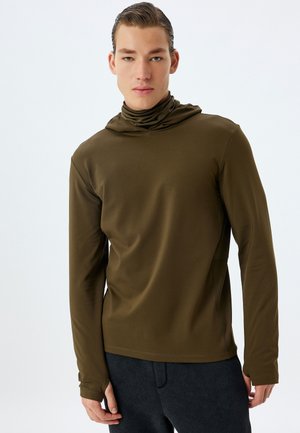 Sudadera con capucha de manga larga en verde oliva, fabricada con un tejido suave y de corte holgado, con detalles de costura. Llevada con pantalones en gris oscuro.