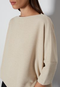 Beige Rippensweater mit einem Bootsausschnitt und dreiviertellangen Ärmeln. Weicher Textur, lockere Passform und dezenter Glanz im Stoff.