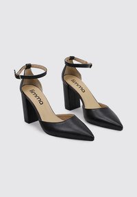 Zapatos de tacón negro con punta afilada, hechos de cuero suave. Cuentan con una correa en el tobillo y un tacón alto grueso. Logo de la marca visible en el interior.