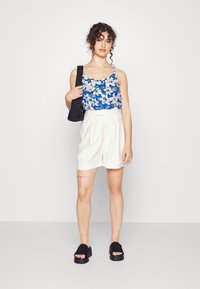 Top de alças azul com flores brancas, calções plissados creme e sandálias plataforma pretas. Modelo segura uma mala de ombro preta.