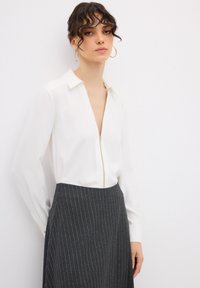 Blusa bianca con zip, colletto e maniche lunghe, abbinata a una gonna a vita alta grigia scura a righe. Tessuto liscio, vestibilità sartoriale.