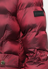 Červená pufovaná bunda s lesklým povrchem, s černou nášivkou "PINKO" a zlatým knoflíkem na přední kapse.