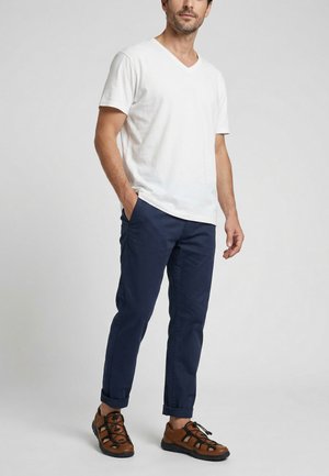 Mann trägt weißes V-Ausschnitt T-Shirt, dunkelblaue hochgekrempelte Hose und braune geschlossene Sandalen, steht mit einer Hand in der Tasche.