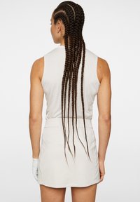 Ljusbeige ärmlös klänning med en tight passform, en dragkedja i ryggen och en kort kjol. Lång flätad hår hänger ner längs ryggen.