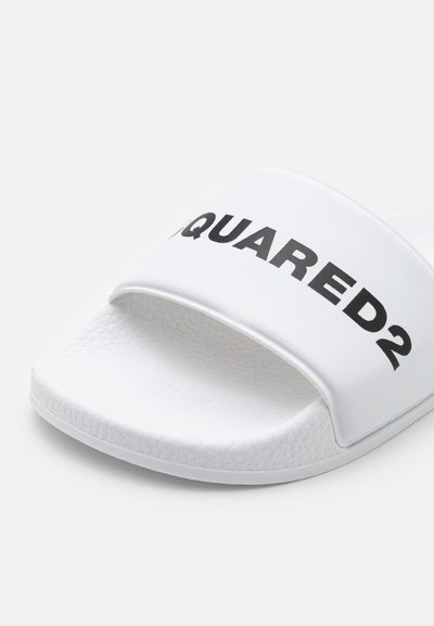 Vit sandaled med texturerad rem som har varumärket "SQUARED2" i stora svarta bokstäver. Platt sula med en slät yta.