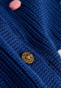 Närbild av blått stickat tyg med en brun knapp märkt "NÄSTA" och en liten rosa texturerad detalj.