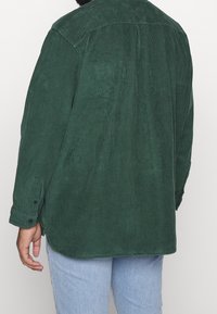 Chemise en velours côtelé vert avec une empiècement dans le dos, manches longues, poignets à boutons et ourlet incurvé. Tissu texturé avec un motif côtelé vertical.