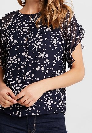 Kobieta ubrana w granatową bluzkę z białymi abstrakcyjnymi plamami, krótkimi falbanowymi rękawami, zapinana na guziki z przodu oraz ciemnoniebieskie dżinsy.