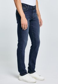 Obcisłe ciemnoniebieskie dżinsy noszone przez osobę w białych trampkach i białej bluzce na jednolitym tle.