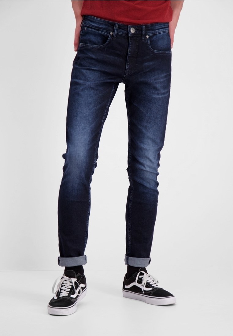Dark blue denim jeans with a slim fit, featuring front pockets, light whiskering, and rolled cuffs, paired with black sneakers.