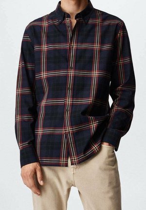 Homme portant une chemise à carreaux bleu foncé, rouge et beige à boutons, avec un pantalon beige, une main dans la poche, sur un fond uni.