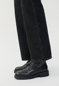 Stivaletti neri in pelle con punta tondeggiante, chiusura con lacci sul davanti e suola testurizzata, abbinati a pantaloni in denim neri larghi.