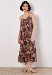 Una giovane donna è in piedi contro uno sfondo chiaro, indossando un vestito maxi a fantasia e sandali beige, con un'espressione neutra.