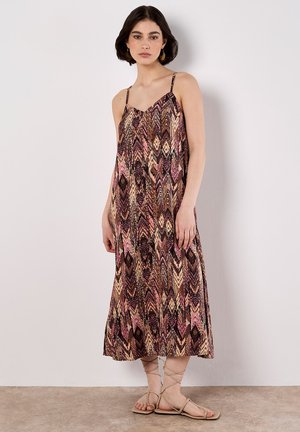 Una giovane donna è in piedi contro uno sfondo chiaro, indossando un vestito maxi a fantasia e sandali beige, con un'espressione neutra.
