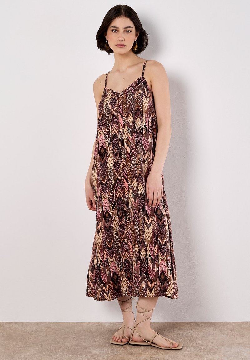 Una giovane donna è in piedi contro uno sfondo chiaro, indossando un vestito maxi a fantasia e sandali beige, con un'espressione neutra.