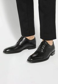 Black leather dress shoes with a polished finish, cap toe design, lace-up closure, and smooth texture, paired with dark trousers.