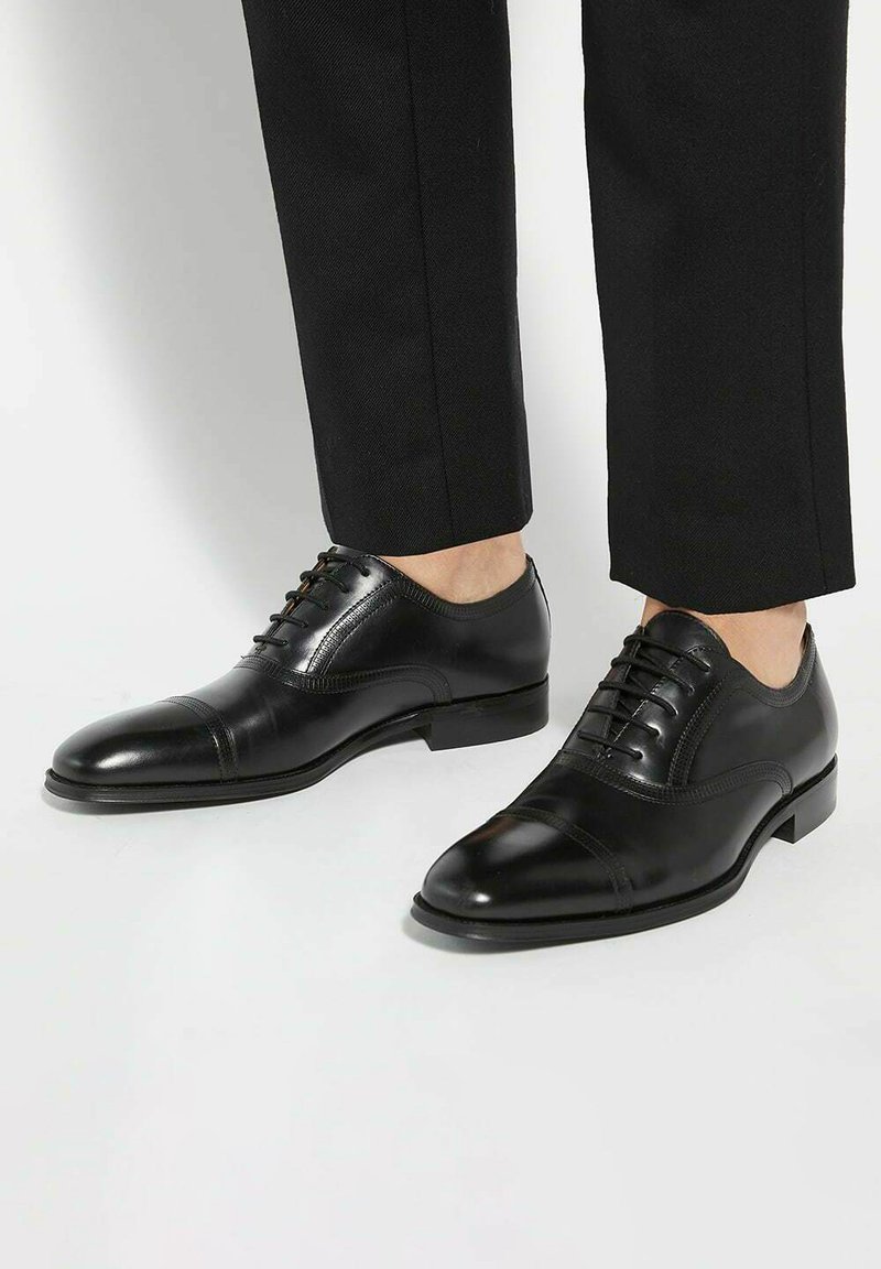 Black leather dress shoes with a polished finish, cap toe design, lace-up closure, and smooth texture, paired with dark trousers.