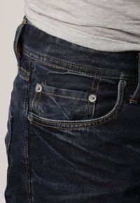 Jeans de denim azul oscuro con costuras visibles y remaches de metal, llevados con una camisa texturizada gris claro metida en la cintura.