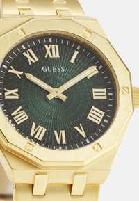 Goudkleurig octagonaal horloge met een textuurrijke donkergroene wijzerplaat, Romeinse cijfers en gouden wijzers. Voorzien van een geschakelde metalen armband.