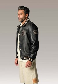 Veste varsity en cuir noir avec des poignets côtelés rayés, des poches avant et un patch brodé. Associée à un pull crème et un short beige.