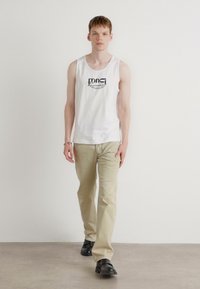Witte tanktop met logo, beige broek en zwarte schoenen. Model staat gecentreerd tegen een effen achtergrond.