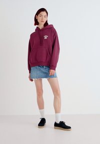 Mörkröd hoodie med framficka och dragsko, ihop med en denimkjol. Svarta skor och vita strumpor kompletterar outfiten.