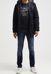 Parka imbottito blu navy con cappuccio foderato in pile grigio, indossato sopra una t-shirt nera con grafica. Abbinato a jeans scuri e scarpe da ginnastica bianche.