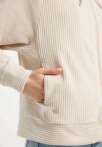 Beige Cord-Jacke mit Reißverschluss, die über eine gerippte Textur, Seitentaschen und ein silbernes Logo-Detail im unteren Taschenbereich verfügt.